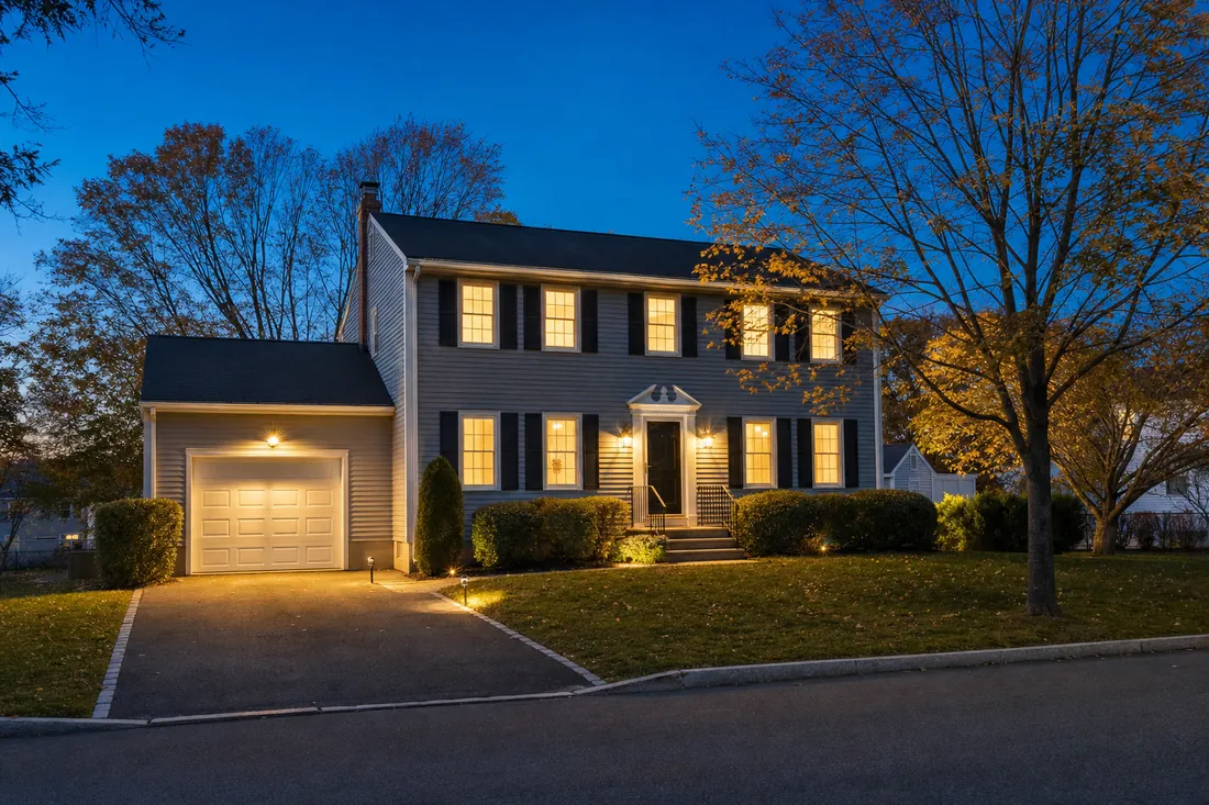 An illustrative example of a two-storey colonial house at twilight, its windows lit warm against a deep blue sky.